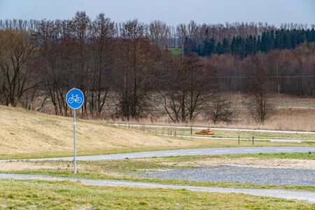 Bicycle path with blue and white road sign in front of the field and forestの写真素材