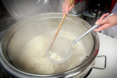 The chef is scooping up cooked noodles from a steaming cauldronの写真素材