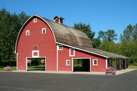 Classic styled red barn with side corral in front of green forested area under blue skiesのeditorial素材