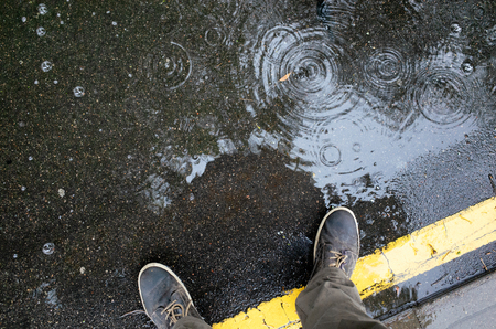 Rainy weather. Male legs in sneakers or boots walking through the rain puddle on the asphalt road, top view. Film grain photo.の写真素材