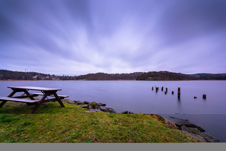 Bench covered with snow next to lake with streams of clouds in the early morning sky and dead logs in the lake gothenburg swedenの写真素材