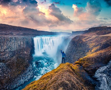 Tourist standing on the shore of falling water of the most powerful waterfall in Europe - Dettifoss. Impressive summer sunrise in Jokulsargljufur National Park, Iceland.の写真素材