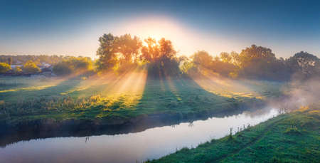Fantastic summer sunrise on the misty river. Panoramic morning scene with first sunlight glowing...