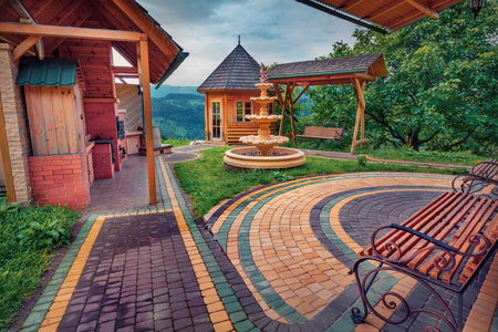 Wooden chalet, decorative fountain and swing on the yard of Carpathian village. Beautiful summer view of mountain village, Ukraine, Europe. Travel concept background.の写真素材