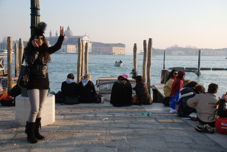 Venice, Italy, 02.21.09: A young woman talks and gestures whilst using her mobile phone during the Venice Carnival. In the background, other carnival goers take a break and admire the famous waterfrontのeditorial素材