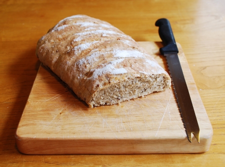 A loaf of home-baked sourdough bread on a wooden chopping board on a wooden table with a large black-handled knife in natural lightingの写真素材