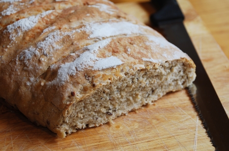 A loaf of home-baked sourdough bread on a wooden chopping board on a wooden table with a large black-handled knife in natural lightingの写真素材