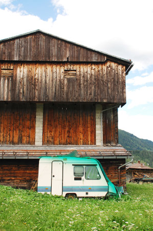 An old disused trailer caravan with a corrugated plastic sheet over its roof in front of an old wooden farming building in north east Italyのeditorial素材