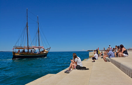 Zadar, Croatia - June 18th 2016. Tourists watch the boats go by as they listen to the Sea Organ (Morske Orgulje) on zadar's waterfont. This experimental musical instrument plays music through the movement of sea waves and tubes positioned under the steps.のeditorial素材