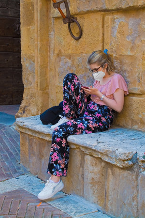 Pienza, Italy - September 6th 2020. A young woman wearing a mask texts in the historic Piazza Pio II in Pienza in Tuscany, Italy, during the COVID-19 pandemic. At this time, mask use is compulsory in the historic centreのeditorial素材