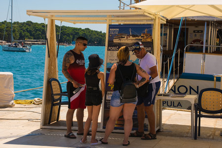 Porec, Croatia- July 10th 2021. A day trip vendor on the seafront tries to sell a boat trip to three passing tourists in the historic medieval coastal town of Porec in Istria, Croatiaのeditorial素材