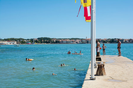 Porec, Croatia- July 10th 2021. Holiday makers and locals use a pier to get into the water at Parentino Beach outside the historic medieval coastal town of Porec in Istria, Croatiaのeditorial素材