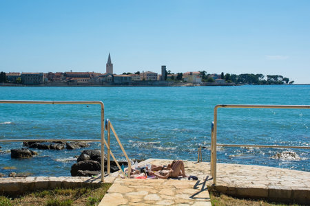 Porec, Croatia - July 9th 2021. Tourists and locals enjoy the summer sun and sea on the coast just north of the historic centre of Porec, seen in the background, on the Istria coast of Croatiaのeditorial素材