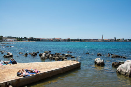 Porec, Croatia - July 10th 2021. Tourists and locals enjoy the summer sun and sea on the coast just north of the historic centre of Porec, seen in the background, on the Istria coast of Croatiaのeditorial素材