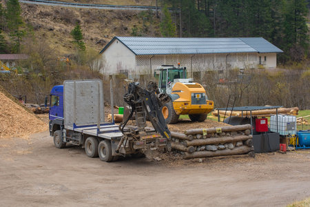 Forni Avoltri, Italy - April 8th 2023. A flatbed container truck with a pneumatic lifting arm and a front end loader tractor at a saw mill yard near the village of Forni Avoltri in Carnia, Udine Province, Friuli-Venezia Giulia, north east Italyのeditorial素材