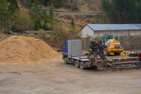 Forni Avoltri, Italy - April 8th 2023. A flatbed container truck with a pneumatic lifting arm and a front end loader tractor at a saw mill yard near the village of Forni Avoltri in Carnia, Udine Province, Friuli-Venezia Giulia, north east Italyのeditorial素材