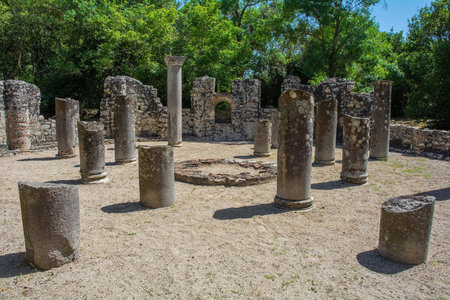 The Baptistry Complex in Butrint Archaeological Park, within Butrint National Park, southern Albania. A UNESCO World Heritage Site. Dating from between 3rd & 6th centuries, this early Byzantine monument features a large circular design with central cross-shaped baptismal font surrounded by 2 concentric colonnades, each with 8 Egyptian granite columns which once supported a wooden roof. The floor is adorned with mosaics covered with sand for preservation. There is a fountain in the background wall. In the centre, an Ionic impost capital featuring acanthus leaves and crossesの写真素材