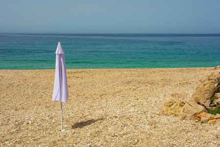 An umbrella on Prinos Beach at the southern end of Himare Bay on the coast of south Albania, part of the Albanian Riviera. A morning in June, early summer, before the sun worshippers have arrived. Located in Vlore County, Himare lies between the Ceraunian Mountains and the Ionian Sea and is a major tourism centreの写真素材
