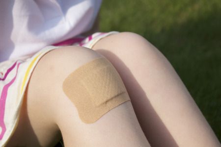 A girl sitting on the grass in a stripey skirt with a big plaster on her knee.  Close up.の写真素材