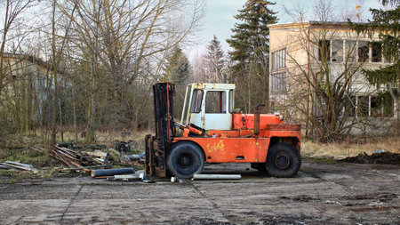 Rusty abandoned forklift sitting in an overgrown lot with scattered debris and a decayed building.の写真素材