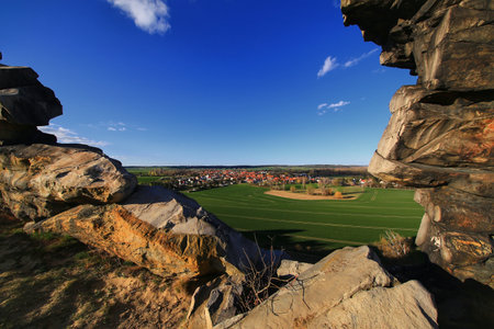 Sandstone frame of the Teufelsmauer, Harz mountains, overlooks village nestled beyond green fields under blue sky.の写真素材