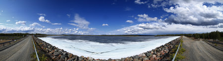 Dam gravel road along icy reservoir edge in northern Sweden under expansive blue sky with scattered clouds.の写真素材
