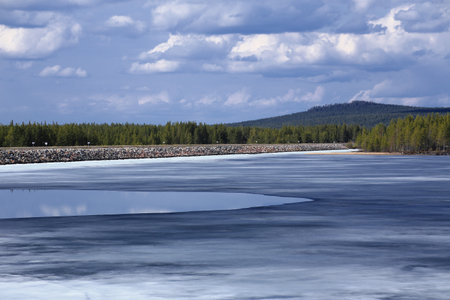 Partially frozen lake bordered by dense forest beneath a cloudy spring sky.