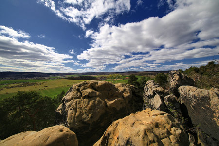 Sandstone cliff edge in the Harz mountains overlooking green fields and hills under a wide blue sky with broken clouds.の写真素材