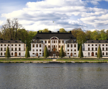White historic Karlbergs palace in Stockholm fronting calm water with flag and trees in a symmetrical landscape.の写真素材