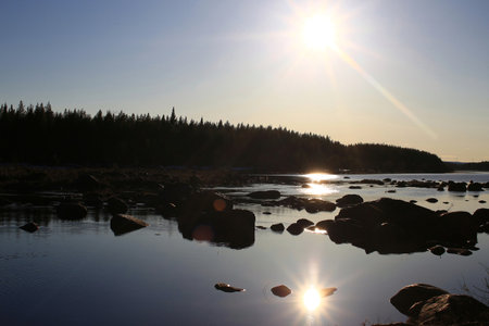 Sunset over rocks along a quiet forest lake with silhouettes and reflected light in water.の写真素材