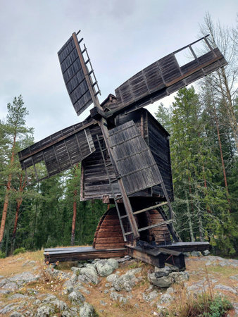 Old wooden windmill in Sweden standing on rocky hill surrounded by pine forest under cloudy sky.の写真素材