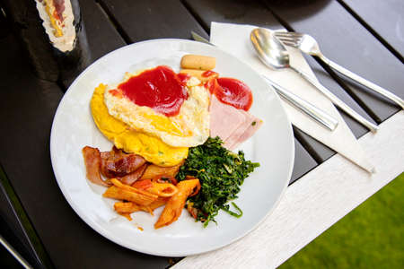 Breakfast from the buffet in a round white plate. on a modern wooden table There is a glass of water upside down, reflecting on it. There are stainless steel knives, spoons, forks placed beside them.の写真素材