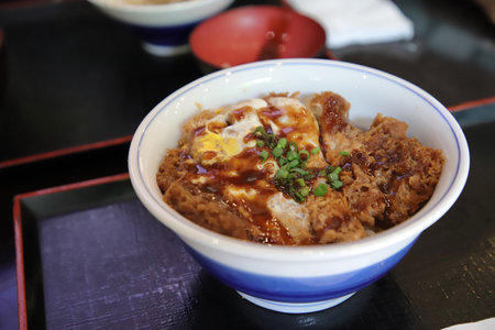 Shallow focus close-up shot of pork and egg on rice with sauce. Japanese food menu Comes in a white tiled bowl on a black wooden tray. The background is a blurred bowl of soup.の写真素材