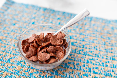 Shallow focus shot of chocolate-flavored breakfast cereal in a clear, circular glass dish on a patterned blue saucer, complete with stainless steel spoons. On a table in a hotel restaurant.の写真素材