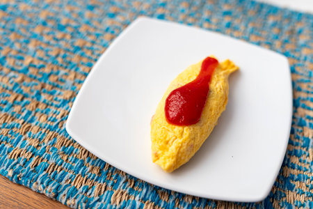A close-up view of a delicious golden yellow omelet. Topped with a little ketchup. Placed on a white plate with a fabric-patterned cloth. The copy area is on the left side of the image. Breakfast.の写真素材