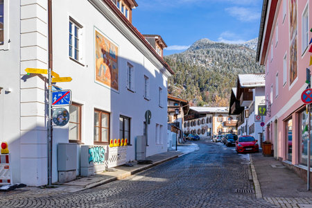 Garmisch-Partenkirchen, Germany â November 28, 2025. A quiet cobblestone street lined with traditional Alpine buildings in the old town of Garmisch-Partenkirchen. A LÃ¼ftlmalerei mural and traffic signs are seen under a clear blue sky, with snow-coveredのeditorial素材