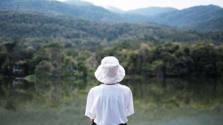 Closeup image of an asian woman white t shirt and hat sitting alone by the river lake with sky and green mountain backgroundの写真素材
