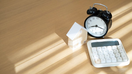 Wooden house calculator and alarm clock on wooden table with sunlight highlighting mortgage deadline...