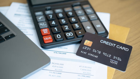 A close up of a black calculator and a black credit card placed on financial documents beside a laptop on a wooden desk symbolizing credit management and financial planning.の写真素材