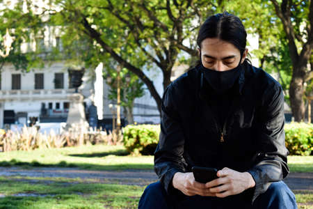 Men looking the phone at the park, with a mask in the middle of a pandemicの写真素材