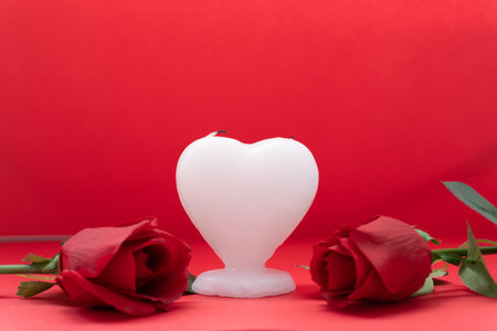 a heart shaped candle with two red roses on each side with a red background, for valentine's dayの写真素材
