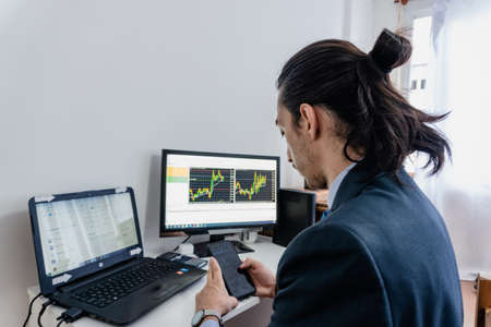 long-haired businessman investing in the stock market, viewing charts on two computers and a cell phone, wearing a blue suit, Labor Dayの写真素材