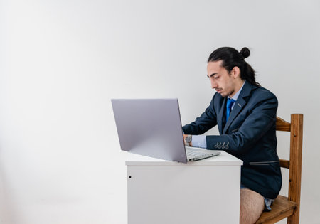 young long-haired businessman working from home with no pants and a monochromatic blue suit, using computer and cell phoneの写真素材