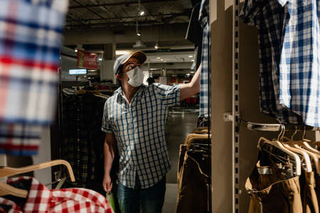man reflected in a clothing store mirror, looking to buy clothes while wearing a medical mask....