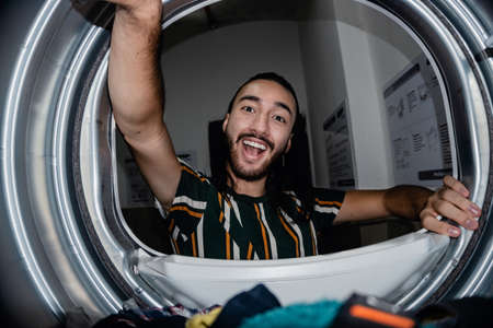 young man with long hair and colorful clothes happily washing in an industrial washing machine. washing at home  the happiness of a quick dry in a tumble dryer.LGBTの写真素材