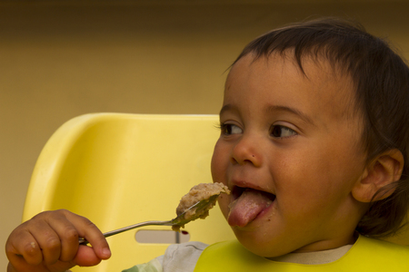 Little happy baby with spoon sits at highchair and eats porridge on plate. Shallow depth of field.の写真素材