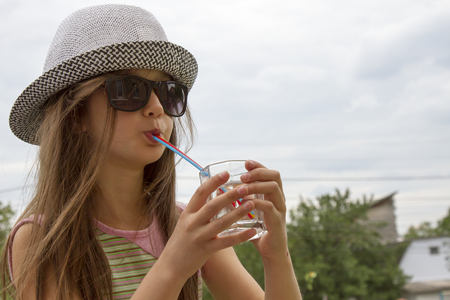 Girl in a hat and glasses drinks water from a glassの写真素材