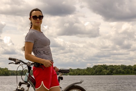 Young woman on a mountain bike stands on the bank of the river