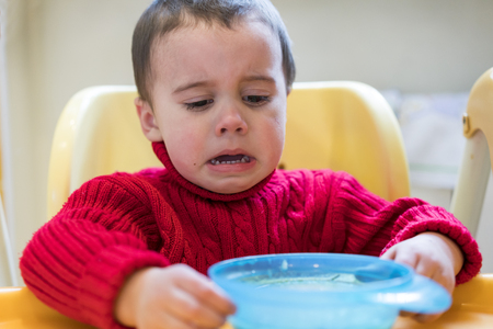 baby boy in red sweater cries. Before him stands a blue plateの写真素材