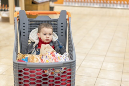 Boy 2 years in a supermarket, sitting in a cart full of various products.の写真素材